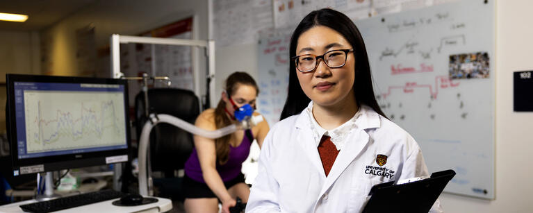 A woman in a white lab coat smiles at the camera while someone else works out behind her