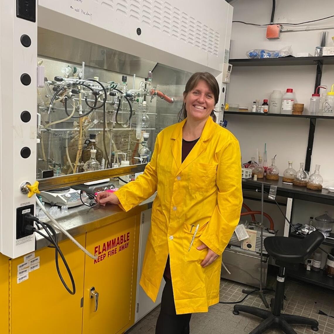 A woman in a yellow lab coat stands in a lab and smiles at the camera