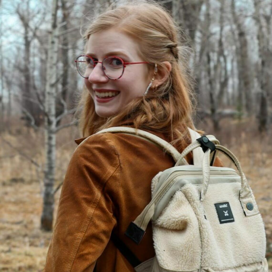 Jess Churcher stands in a forest wearing a backpack and a brown coat and smiles back at the camera