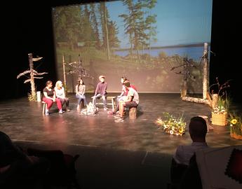 People on a stage sitting around an unlit campfire (ii’ taa’poh’to’p Indigenous UCalgary)