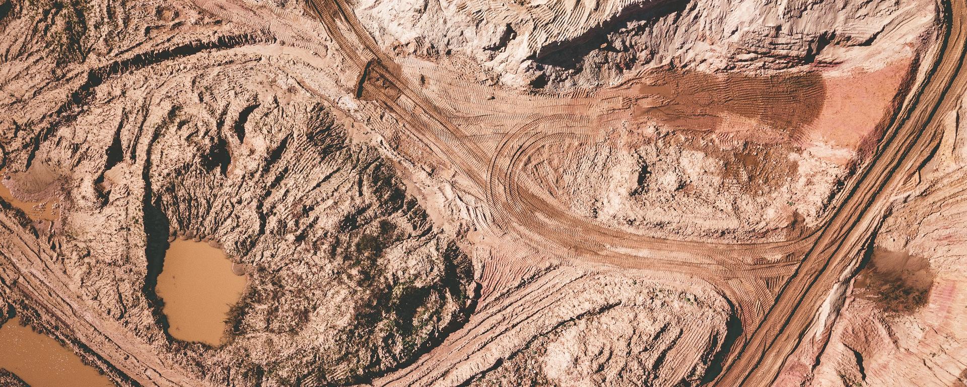 An overhead view of an open pit mine. The ground is brown and rusty-looking, and there are dirt roads leading into and out of the mining area.