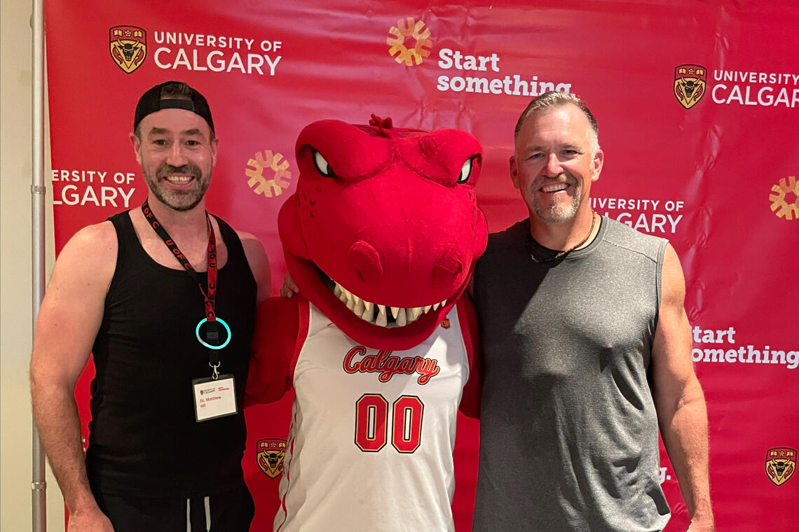 two men in workout attire posing with a dinosaur mascot at a spin-a-thon event