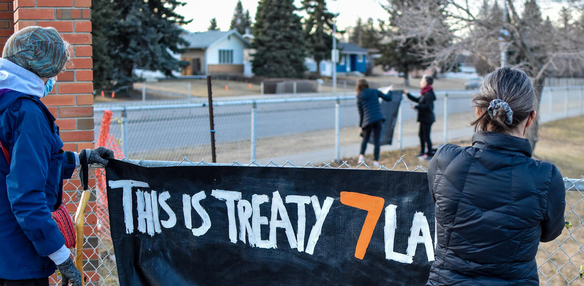 A person in a blue coat and a person in black coat hold up a black banner that says "this is Treaty 7 land" in white and orange letters.