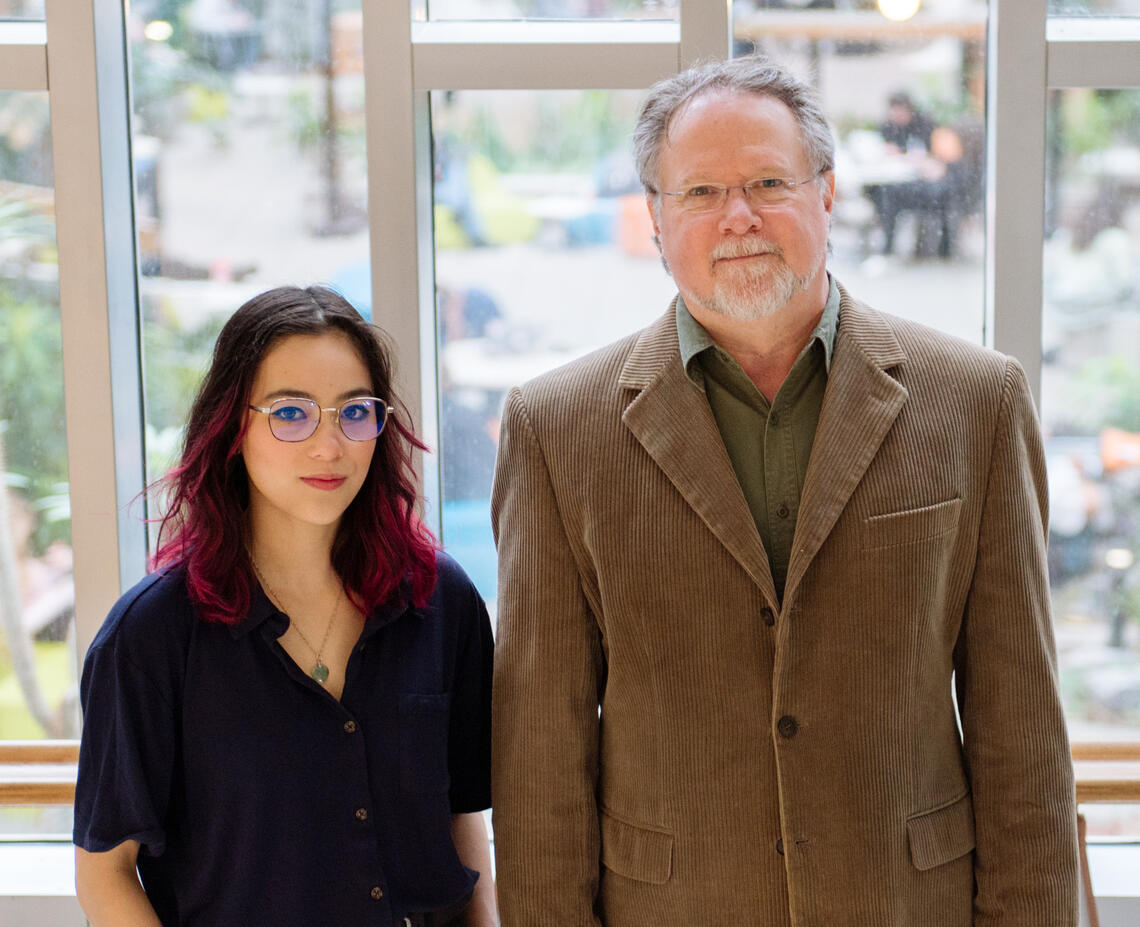 Emilie Lui, a student wearing glasses with dark hair and pink ends, stands beside Paul Stortz, a professor with grey hair and glasses wearing a blazer. They are standing inside with a glass wall into an atrium behind them.