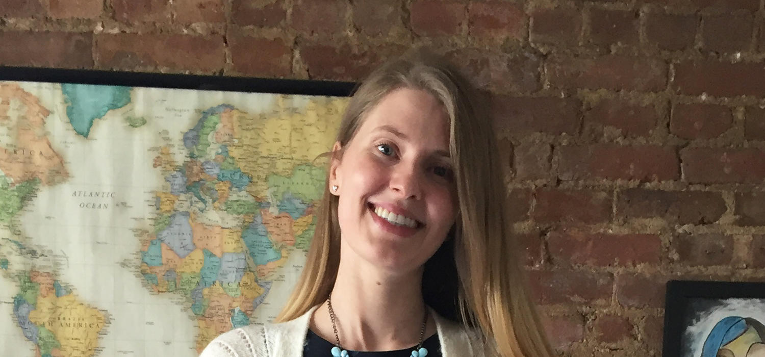 Alumna Alexandra Kennedy smiles in front of a brick wall with a map poster on it