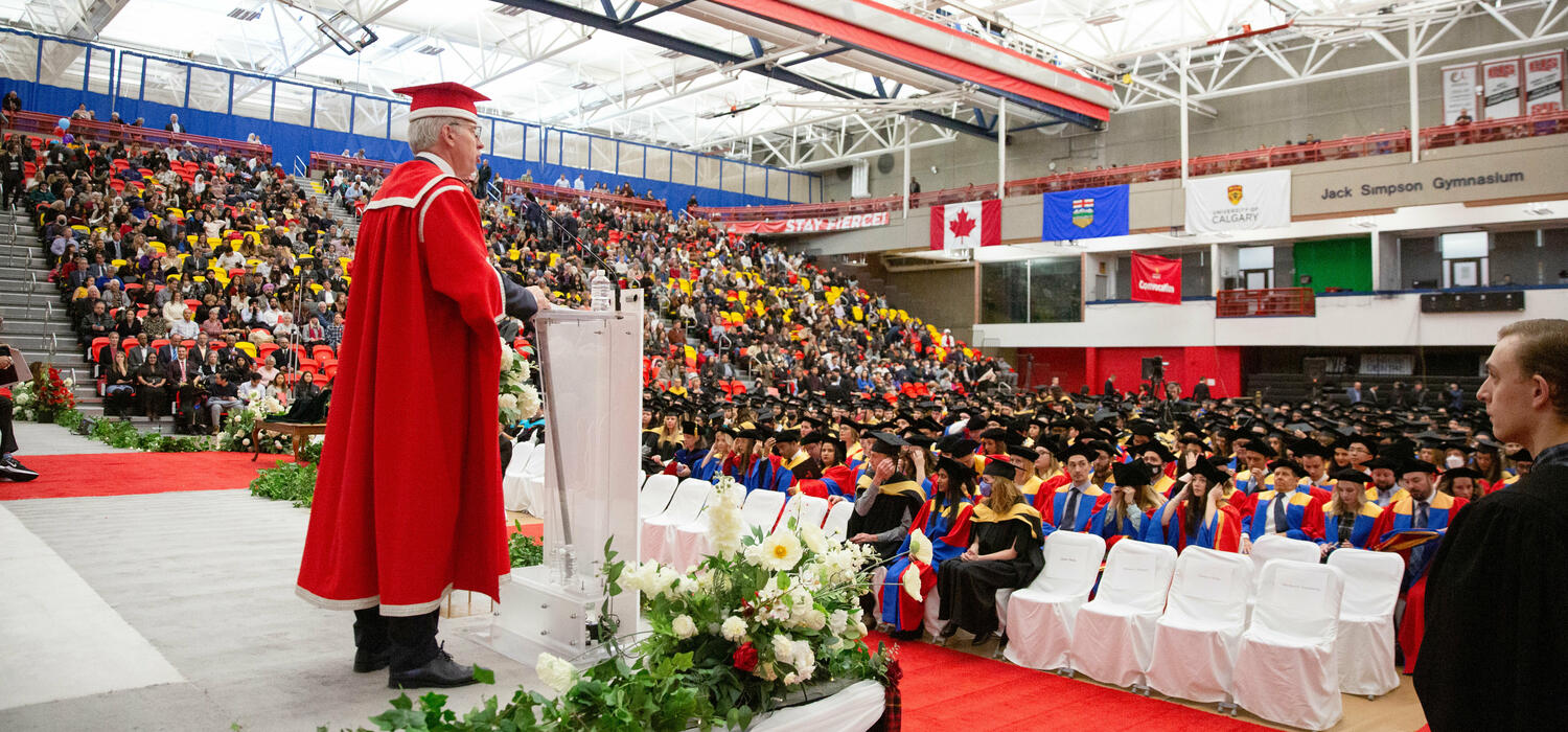 A shot from convocation of a man in a cap and gown on a stage looking out to a gym full of graduates