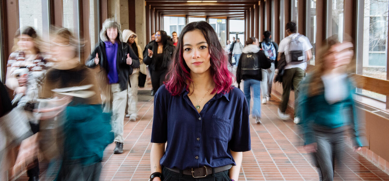 Emilie Lui, a young student with dark hair that has pink on the ends, stands in a busy hallway with blurred people walking around her. They are facing forward and have their hands in their pockets.