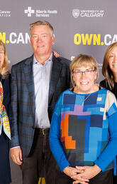 4 people stand next to each other smiling at the camera in from of a banner that has University of Calgary, Alberta Cancer Foundation, Alberta Health Services and OWN.CANCER written on it