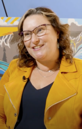 A woman in a yellow jacket smiles with artwork behind her