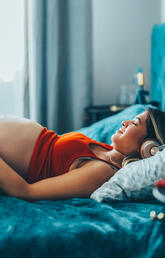 Pregnant woman wearing headphones laying on a bed.