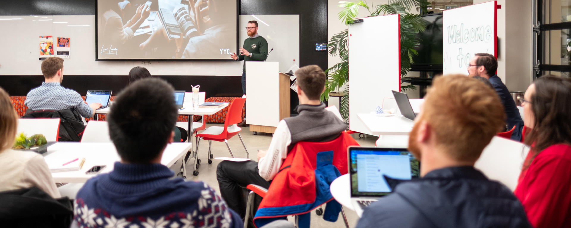 Students sitting at desks in a classroom looking at the front of the room where someone is giving a presentation