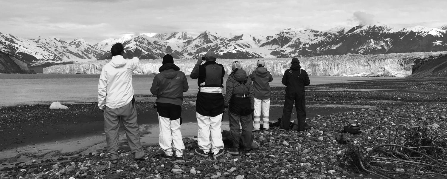 Six geoscientists stand on a cobble beach looking to the North at the Hubbard Glacier where it enters Disenchantment Bay in Alaska