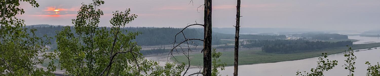 view overlooking a river at sunset with a cloudy sky, red sun in the background and green bushes in the foreground