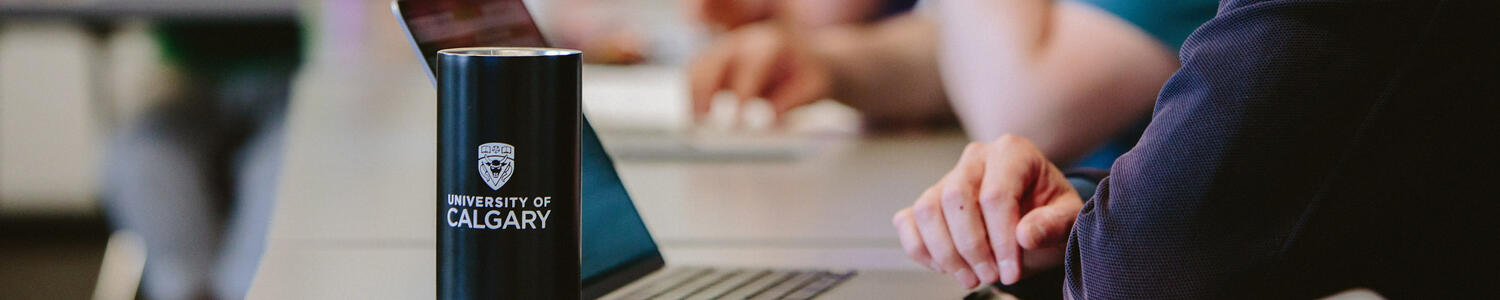 A University of Calgary mug beside a laptop with a meeting in the background.