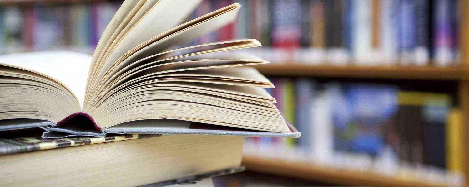 Image of a stack of books with one book opened on top and library shelves behind