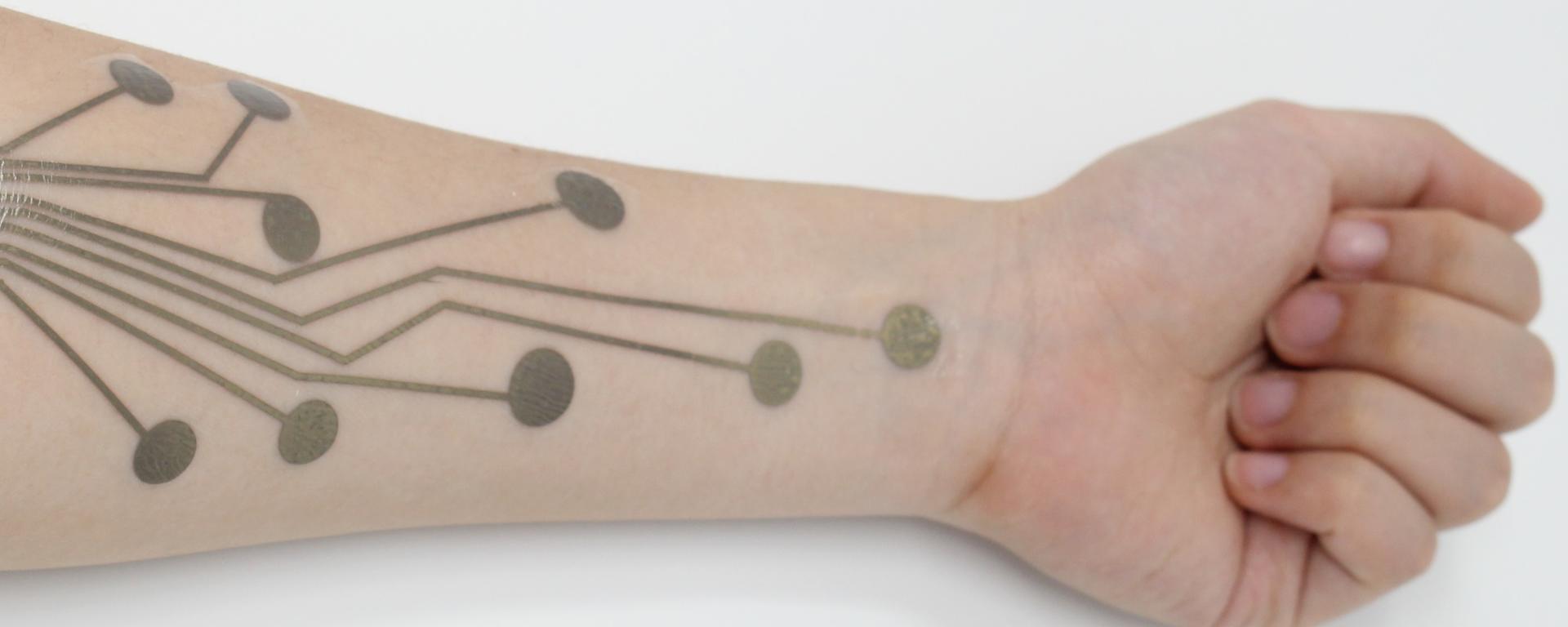 PhysioSkin temporary tattoo device on the forearm. The device has printed electrodes for physiological sensing.