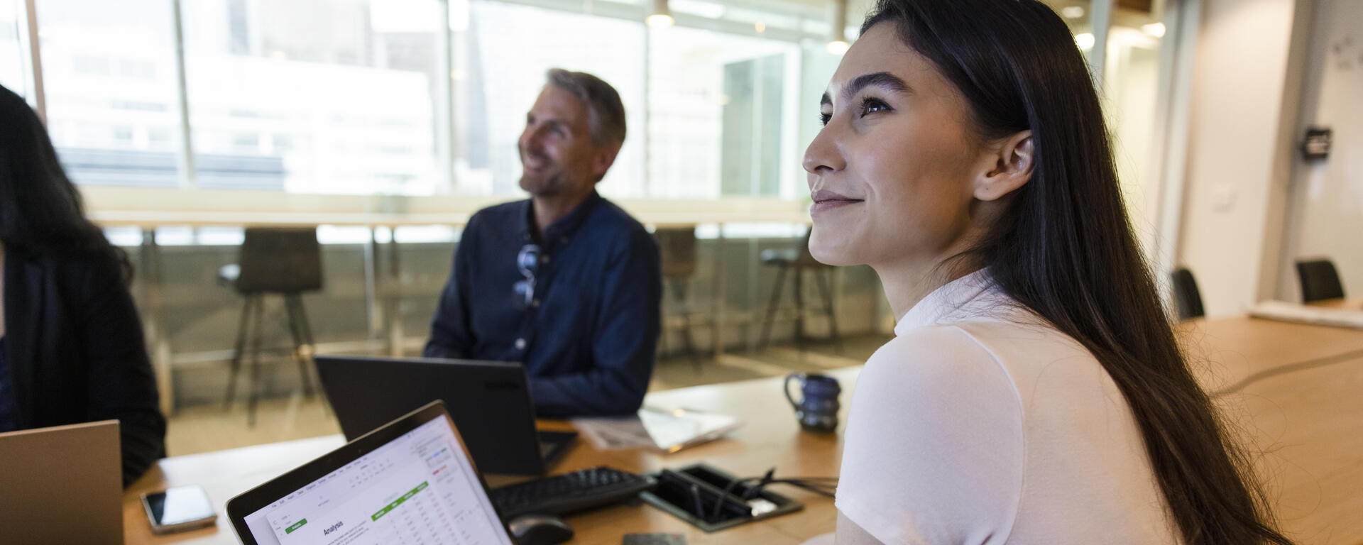 A young woman on a laptop smiling with a colleague smiling in the background.