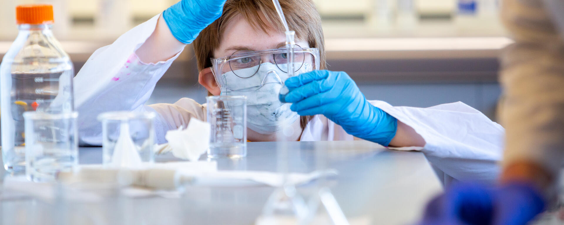 A student in a lab wearing safety glasses, mask and gloves uses a syringe to drop liquid into another vessel.