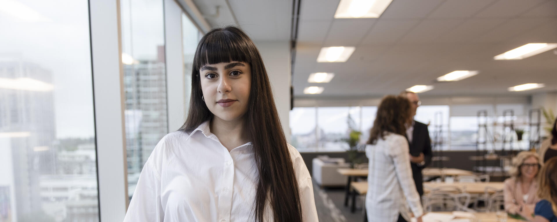 A woman with long dark hair and bangs stands in an office by a window looking straight ahead with a slight smile. She is wearing a button up shirt and striped pants and holding a folder.