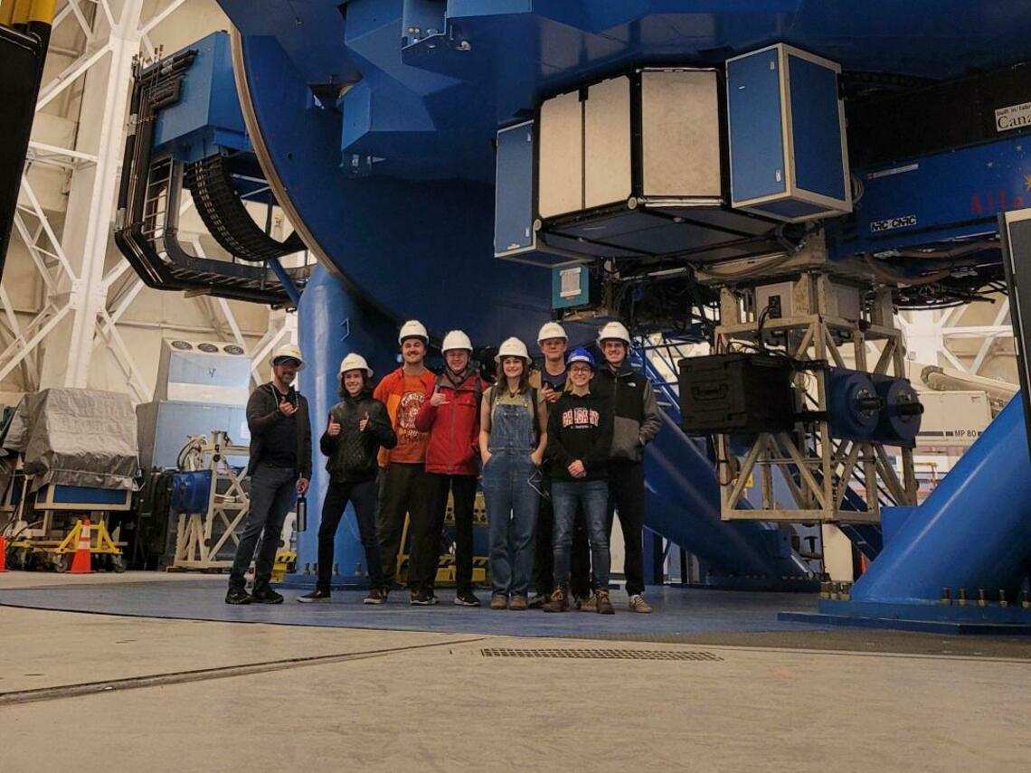 Group of science students in hard hats smiling at the camera while standing in front of large equipment.