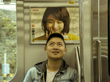 Image of a young man sitting on a train and looking up at a poster of a woman looking down at him