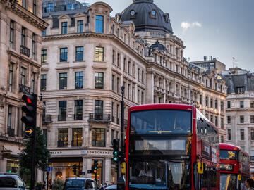 This portrait captures the London street, old beige buildings along the street and a bright red transit bus coming towards the camera. 