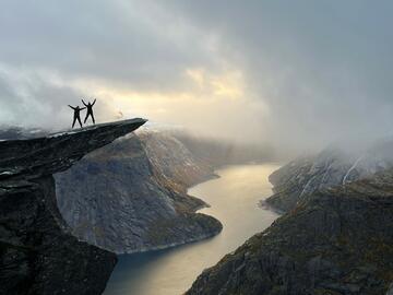 A long distance shot of two silhouetted figures standing at the edge of a cliff. The cliffs overlook a misty river that runs through them.