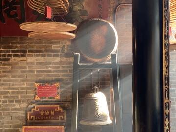 A red-bricked room, where sunlight streams in from the right side. There are Chinese wooden lanterns hanging from the ceiling and a large bell and gong stood at the far end of the photo. 