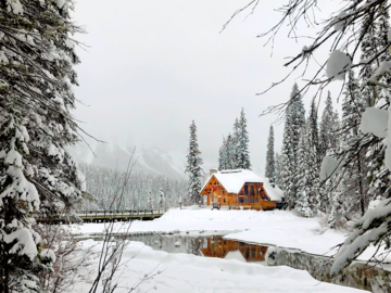 This photo is a snow scape framed with snow-crusted trees on either side of the foreground and all along the background. On the lower third right corner of the photo is a little orange lodge standing out among the white landscape.
