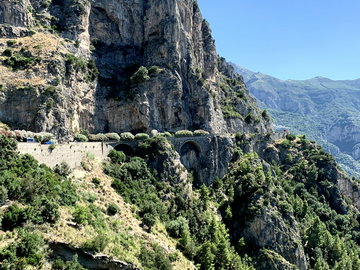 A hill road runs across a mountain in frame. The background features more mountains shouldered behind the one in focus. Blue skies, green hills, and a rocky ridge. 