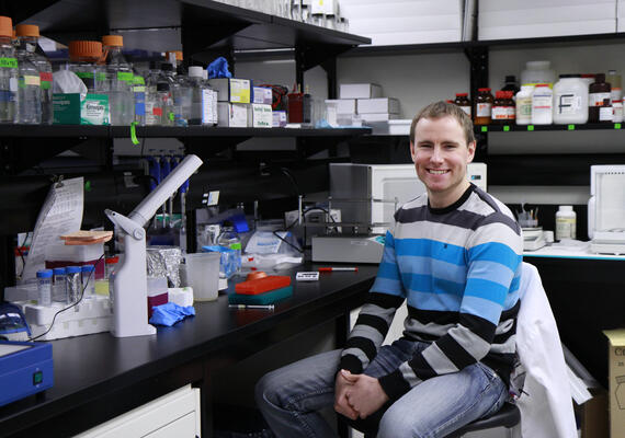 A young researcher sits in a lab and smiles.