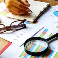 A magnifying glass, a pair of glasses, and a hand writing on a notepad, all on top of a table covered with papers with charts and graphs on them