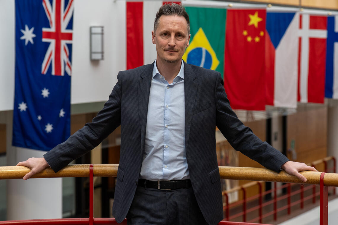 A man wearing a suit stands on a balcony in front of various flags