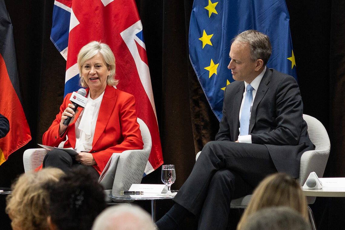 Two people sit on a panel in front of multiple nations flags while speaking into microphones